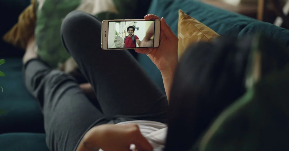 Une femme se détend sur un canapé tout en vidéo discutant sur son smartphone à l'intérieur.
