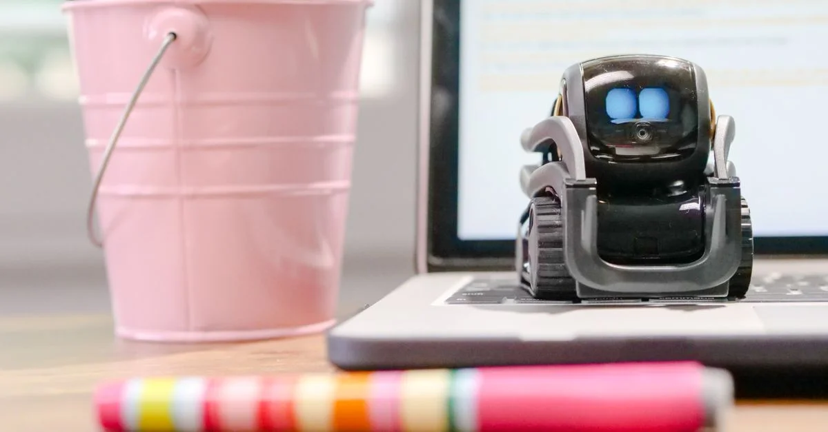 Un petit robot est assis sur le clavier d'un ordinateur portable à côté d'un porte-stylo rose, symbolisant la technologie et l'innovation.